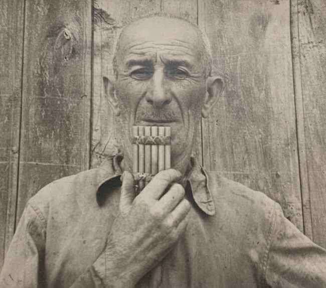 Sepia photograph of an elderly man in a collarless shirt, standing against weathered boards, holding a soinari to his lips with both hands.